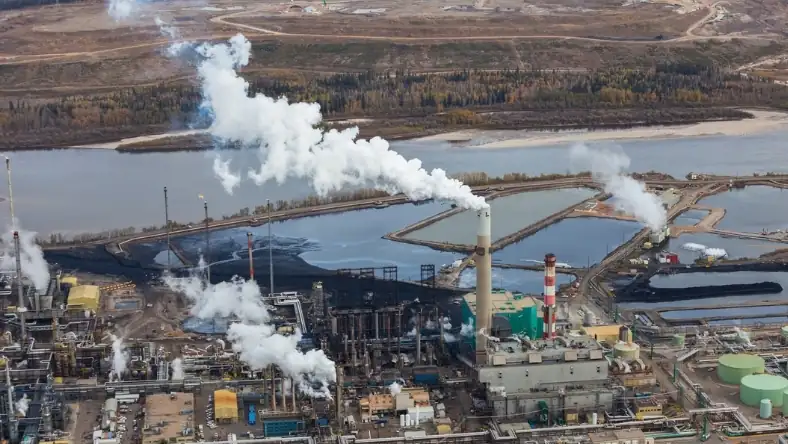 Aerial view of an oil sands facility with steam stacks and a dark tailings pond beside a forested river