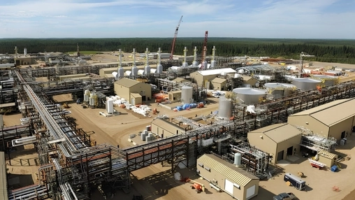 Aerial view of an oil sands processing facility with pipelines, tanks, and cranes surrounded by boreal forest